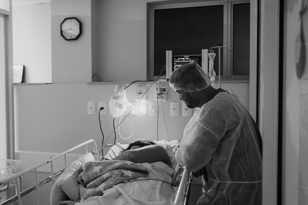 “Pai observa com ternura a mãe no leito hospitalar no pós‑parto em cena em preto e branco”