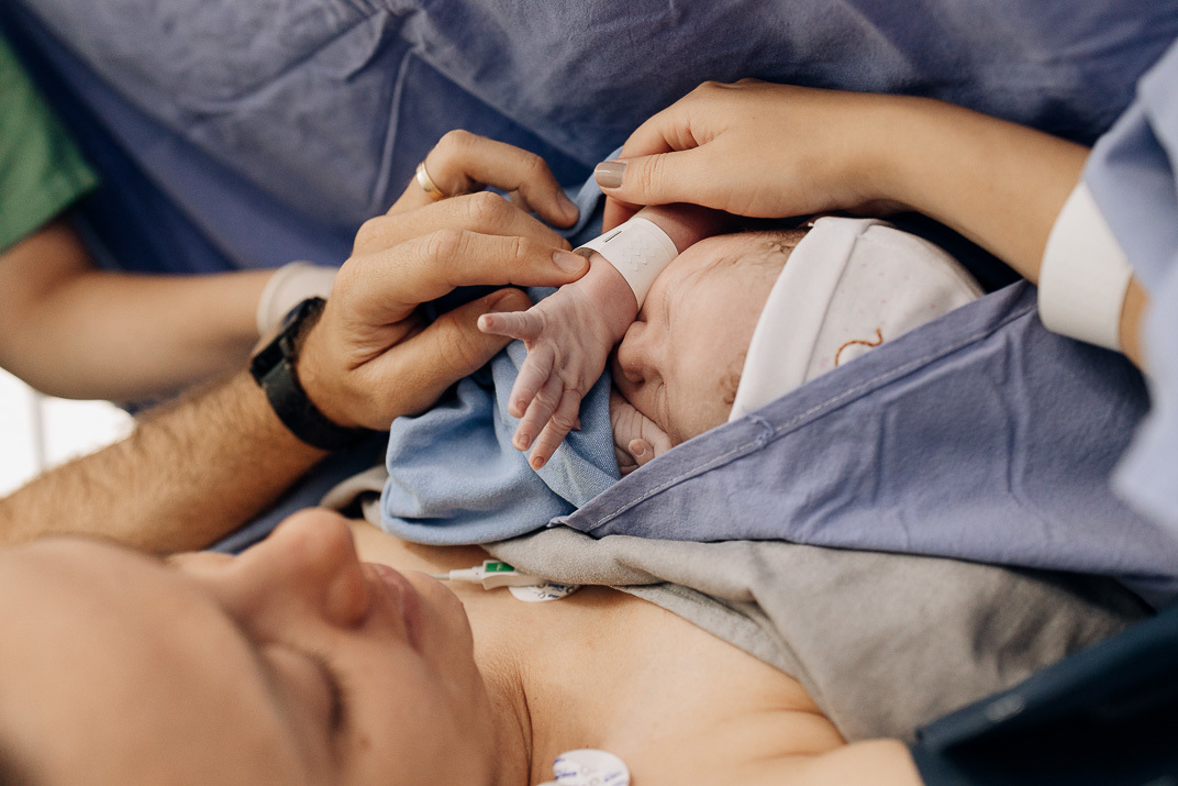 Recém‑nascido no colo da mãe com mão protegida por enfermeira e toque do pai no Hospital Bruno Born