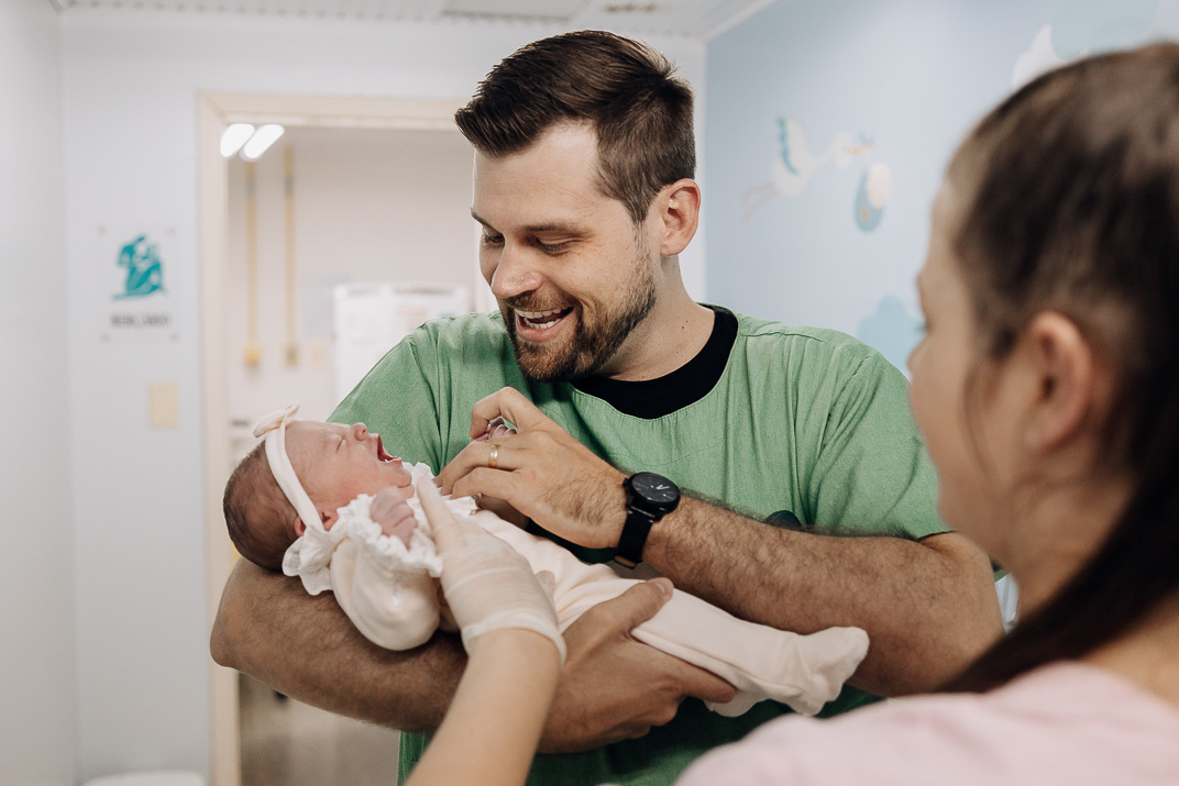 Pai sorri enquanto segura o bebê que chora levemente, com mãe ao fundo, no Hospital Bruno Born