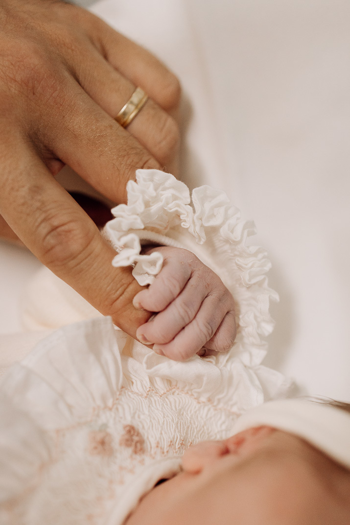 Pequena mão do bebê segurando o dedo do pai, com delicada luva branca sobre o punho do recém‑nascido