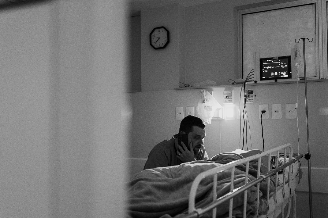 Pai sorri enquanto segura o bebê que chora levemente, com mãe ao fundo, no Hospital Bruno Born