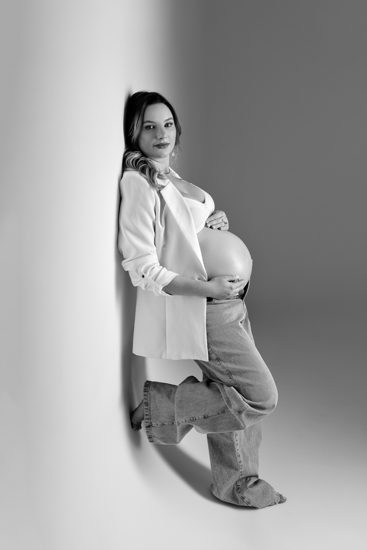 Gestante em PB apoiada na parede, blazer branco e sutiã, calça jeans, mãos envolvendo a barriga; luz suave de estúdio