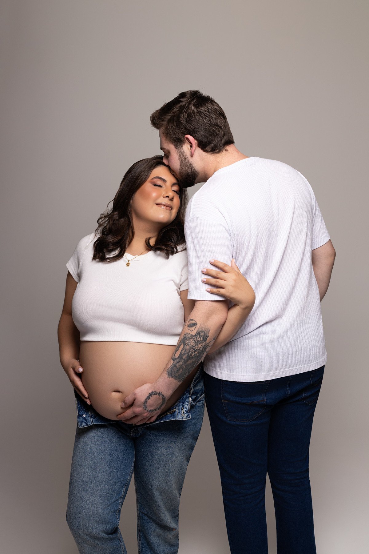 Casal em estúdio: parceiro beija a testa da gestante, mãos do casal apoiadas na barriga; clima de carinho e intimidade