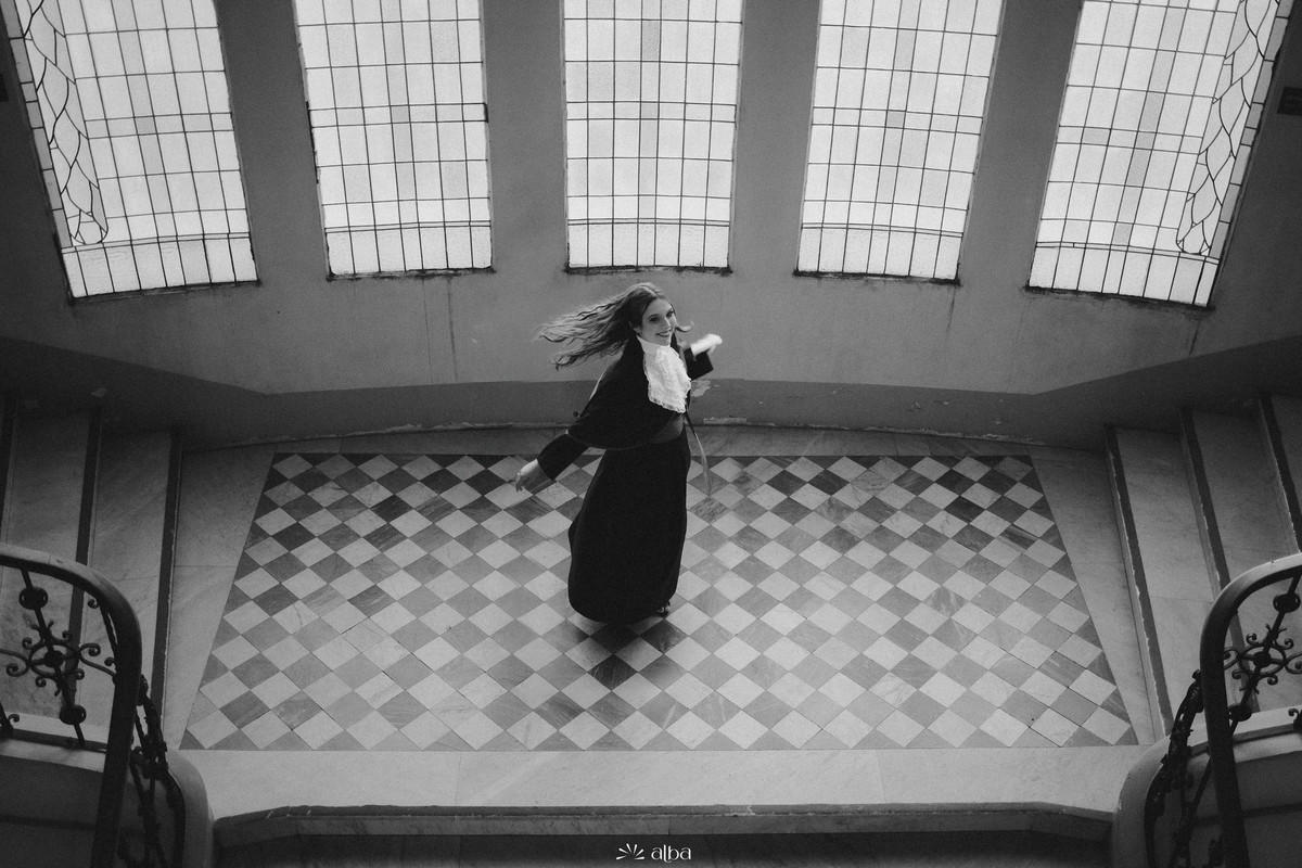 Foto em preto e branco de formando girando com a beca sob vitrais e piso xadrez no prédio da UFRGS.