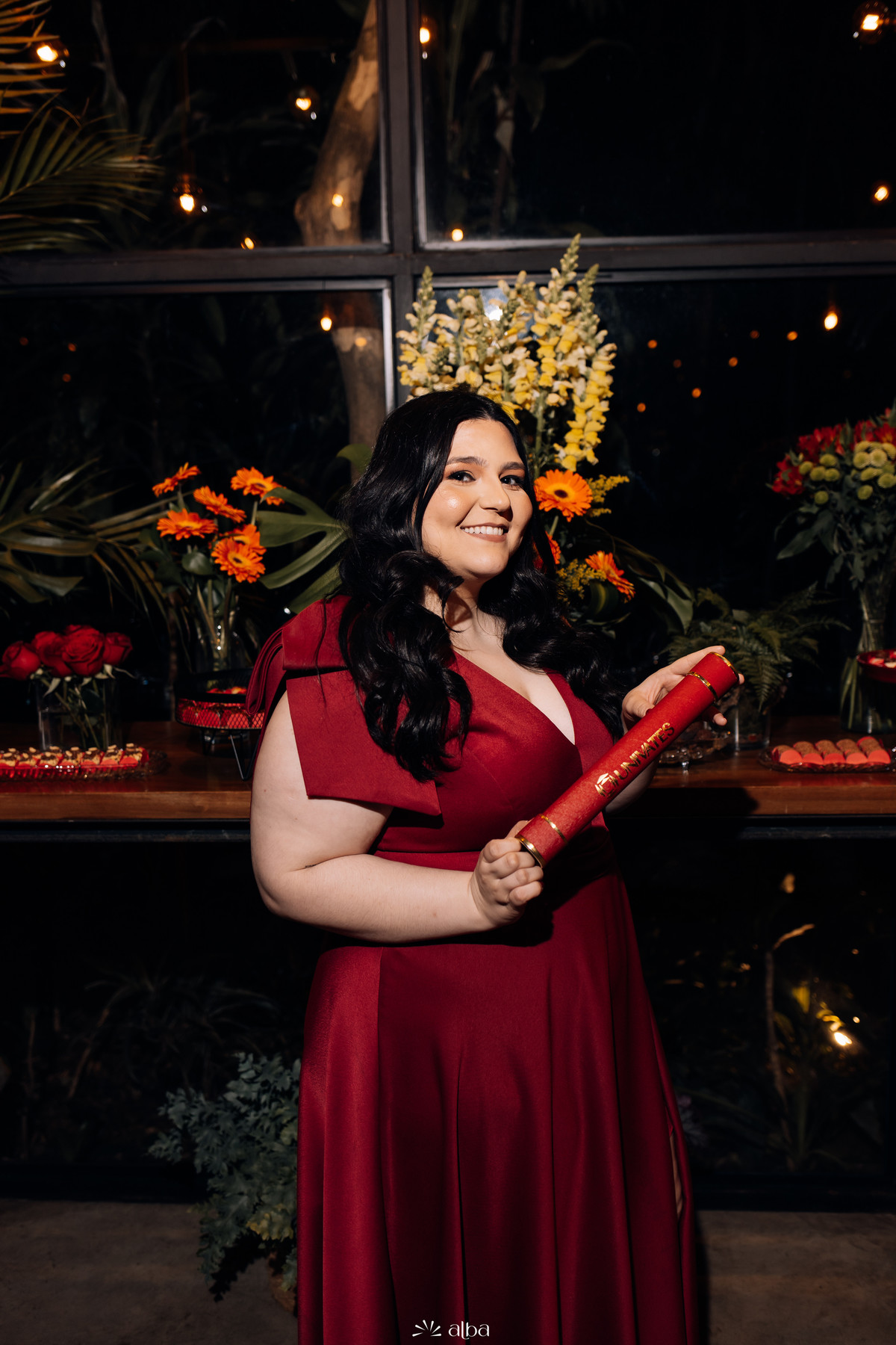 Retrato da formanda com o canudo junto à mesa de doces e arranjos florais.