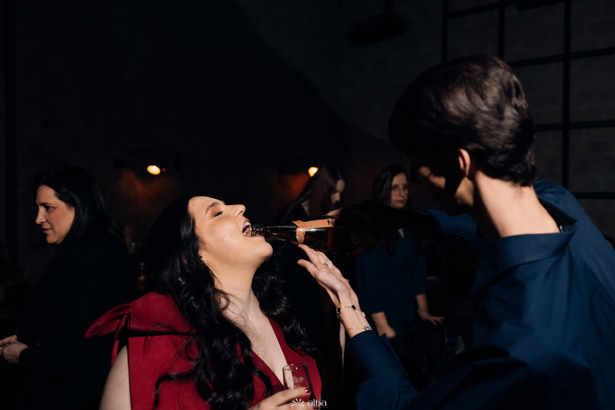 Amigo servindo espumante diretamente na boca da formanda; clima de celebração.