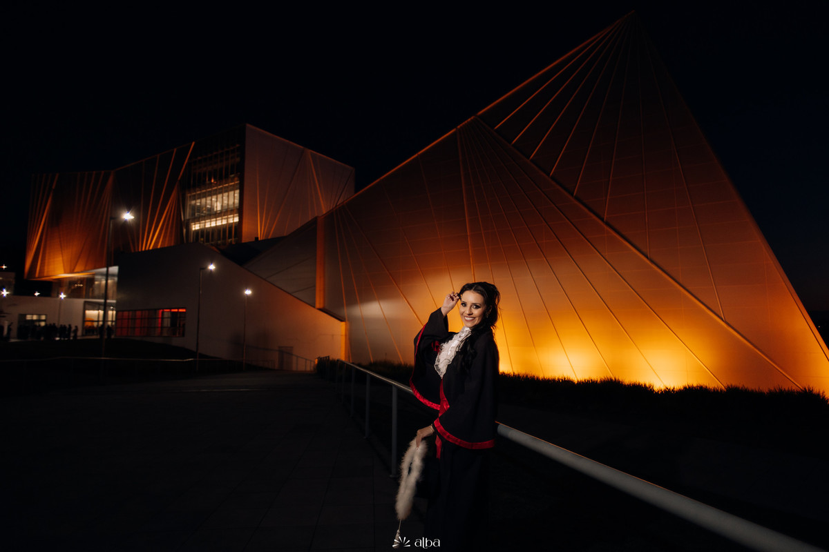 Lígia de toga em frente à arquitetura iluminada da Univates à noite, sorrindo e segurando o capelo com plumas.