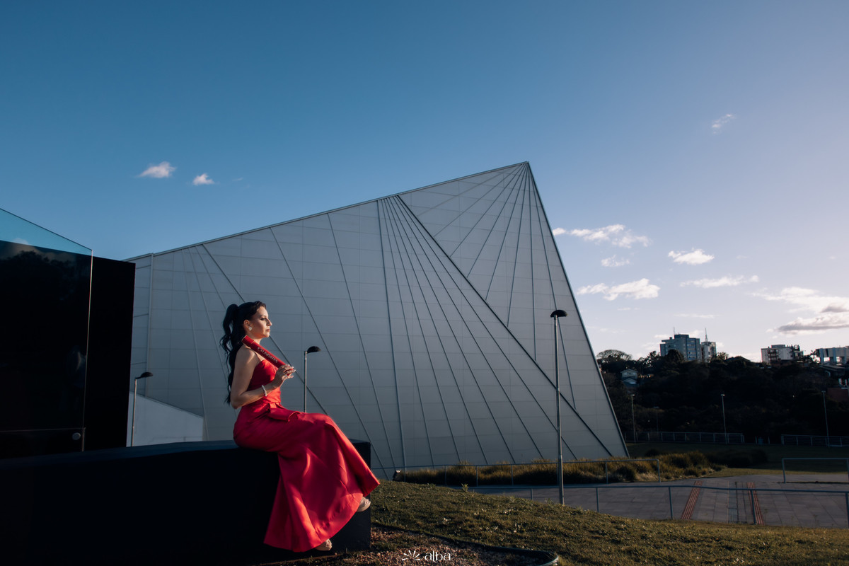 Foto ampla com Lígia sentada observando o prédio geométrico da Univates ao entardecer.
