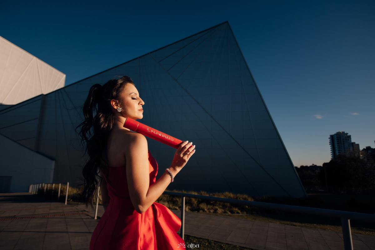 Close de Lígia com canudo apoiado no ombro, luz dourada e prédio da Univates ao fundo.