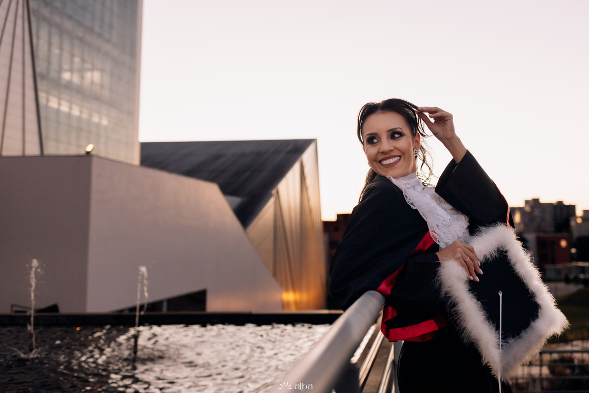 Lígia de toga na passarela da Univates ao entardecer, água da fonte ao fundo.