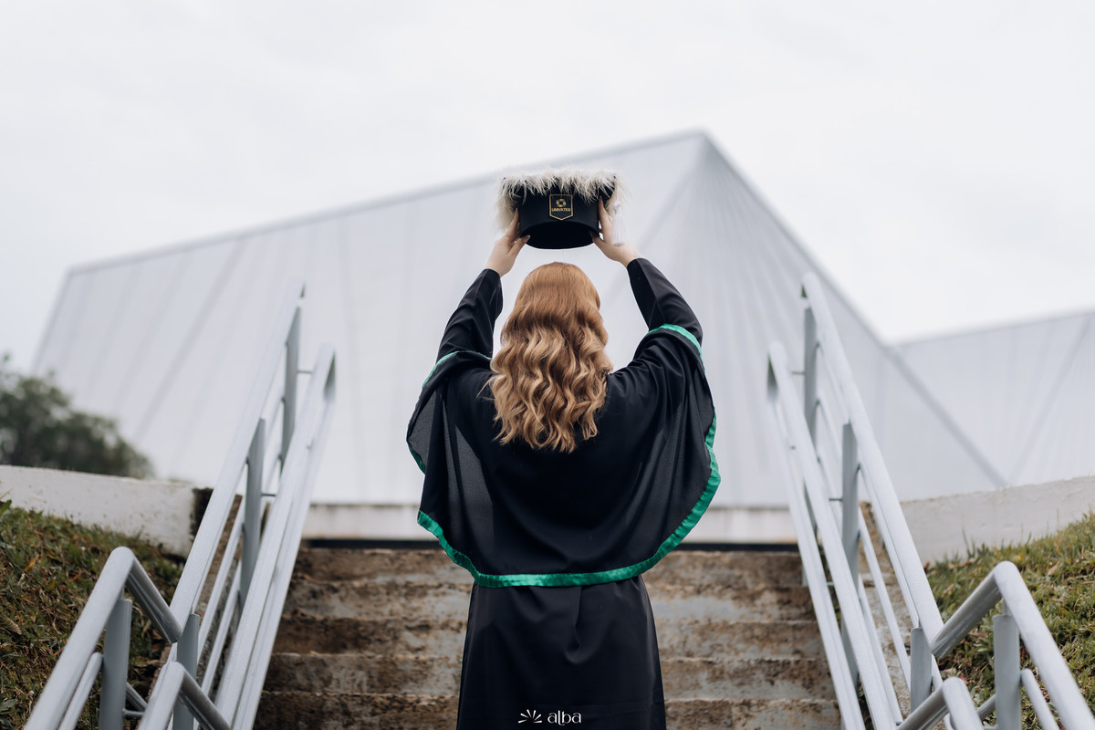Foto de costas da formanda levantando o capelo em frente ao prédio da Univates – Alba Estúdio, Lajeado.