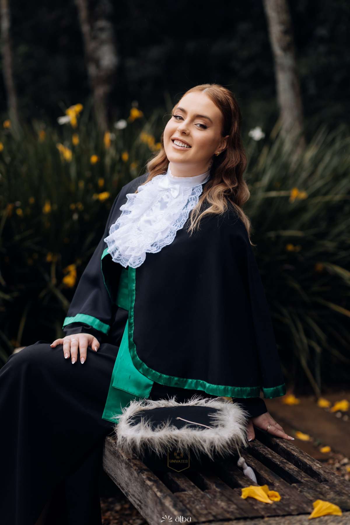 Formanda sentada em banco de madeira no ensaio de toga, segurando capelo com plumas – Alba Estúdio, Lajeado.
