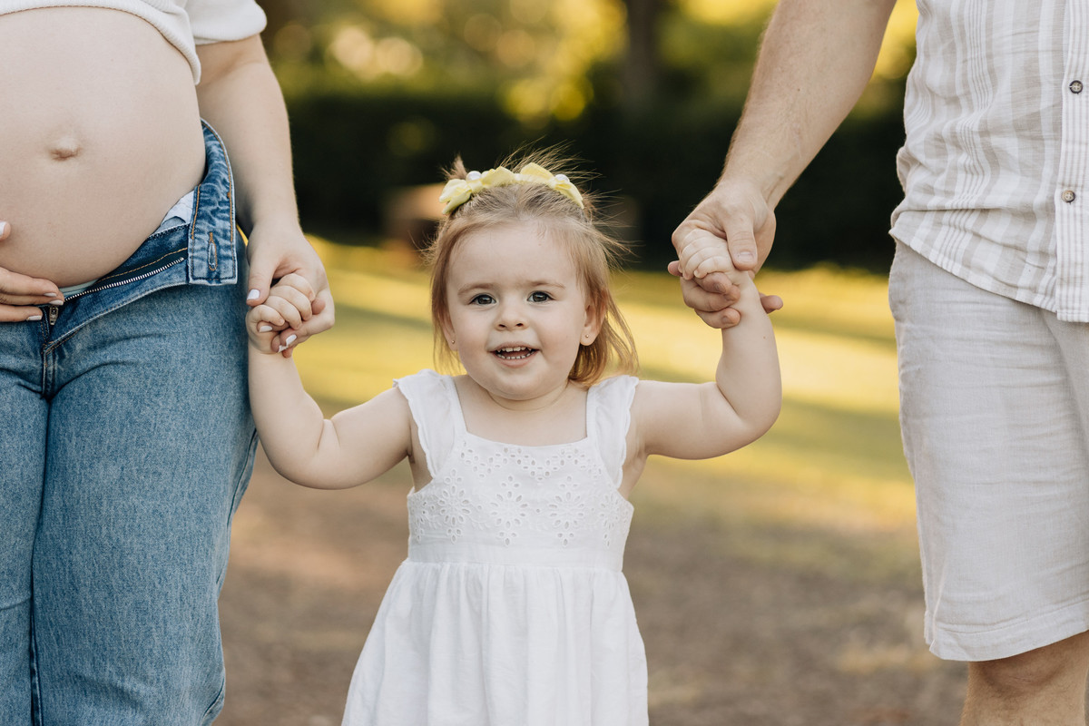 Menina sorrindo entre os pais, segurando as mãos deles, em ensaio gestante ao ar livre em Lajeado – fotografia por Alba Estúdio.