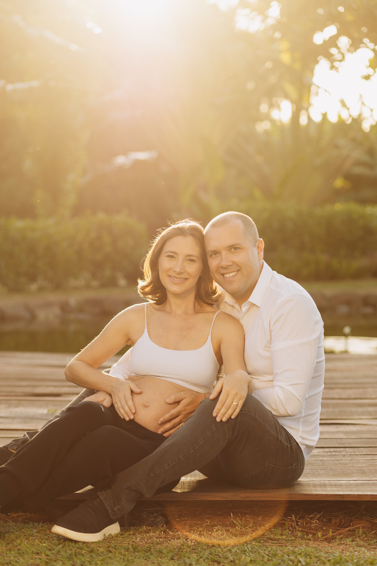 Casal sentado em passarela de madeira ao pôr do sol, trocando olhares carinhosos durante ensaio gestante em Lajeado – fotografia por Alba Estúdio.