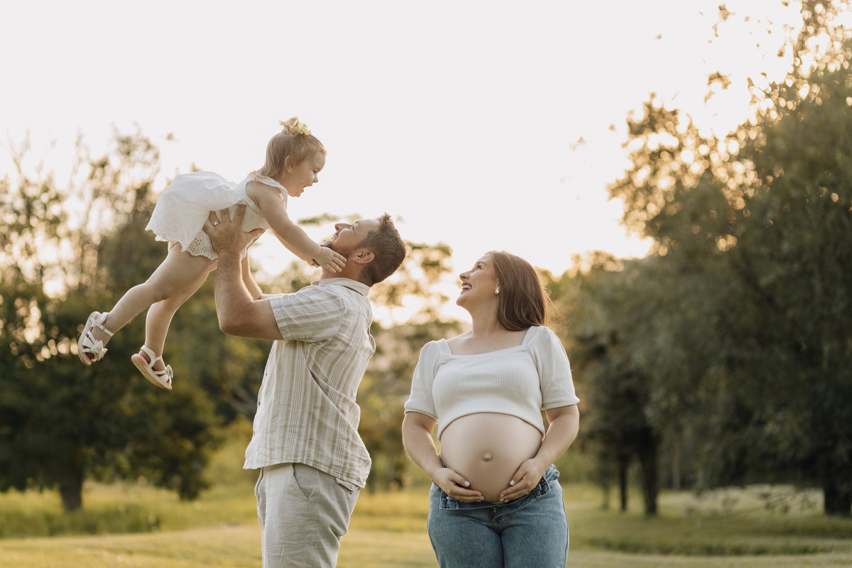 Pai levantando a filha no ar enquanto a mãe gestante observa sorrindo, em ensaio externo cheio de amor e luz natural – fotografia por Alba Estúdio.