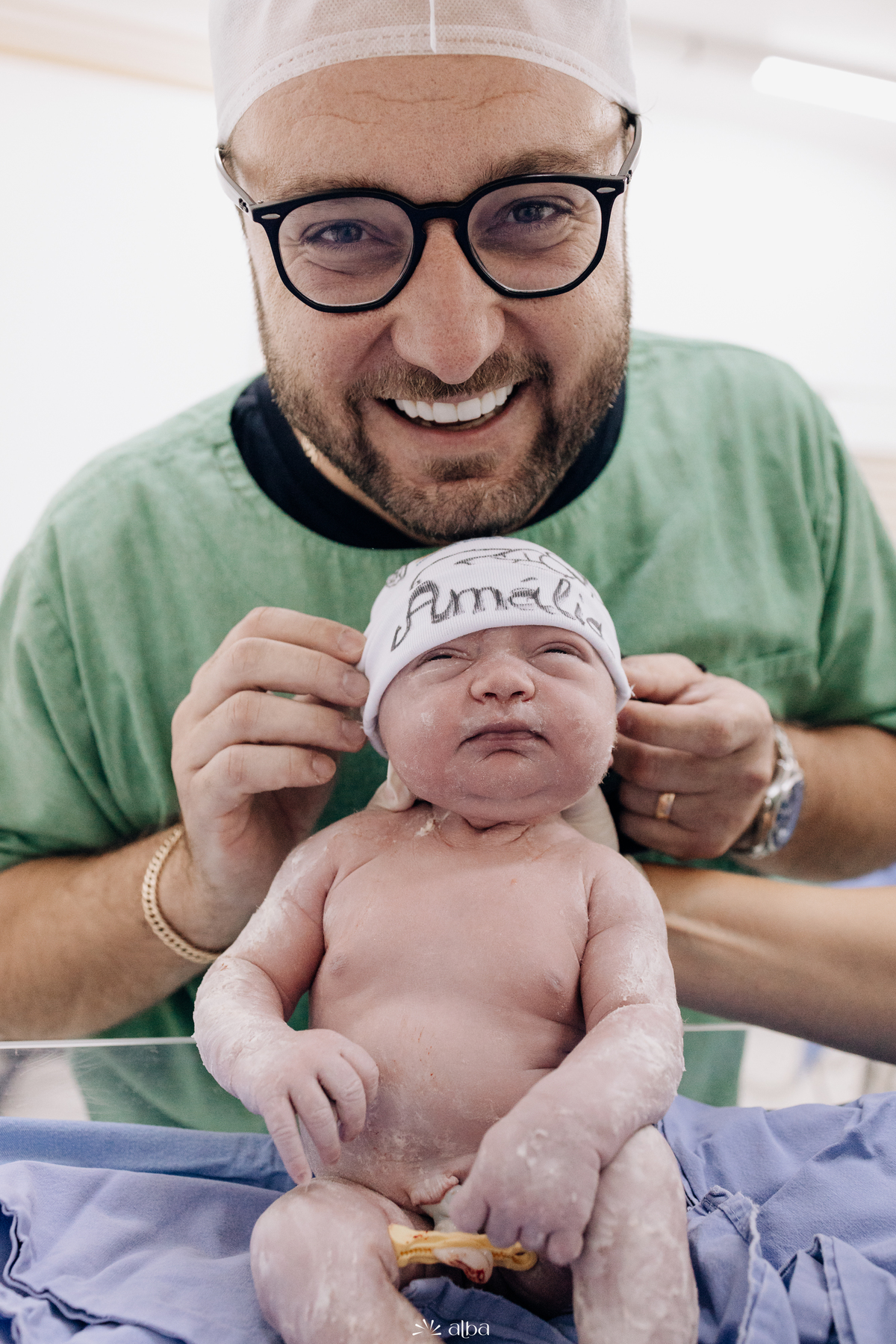 Pai segura a recém-nascida Amália com gorro personalizado na sala de parto do Hospital Bruno Born, Lajeado/RS — foto feita pelo Alba Estúdio.
