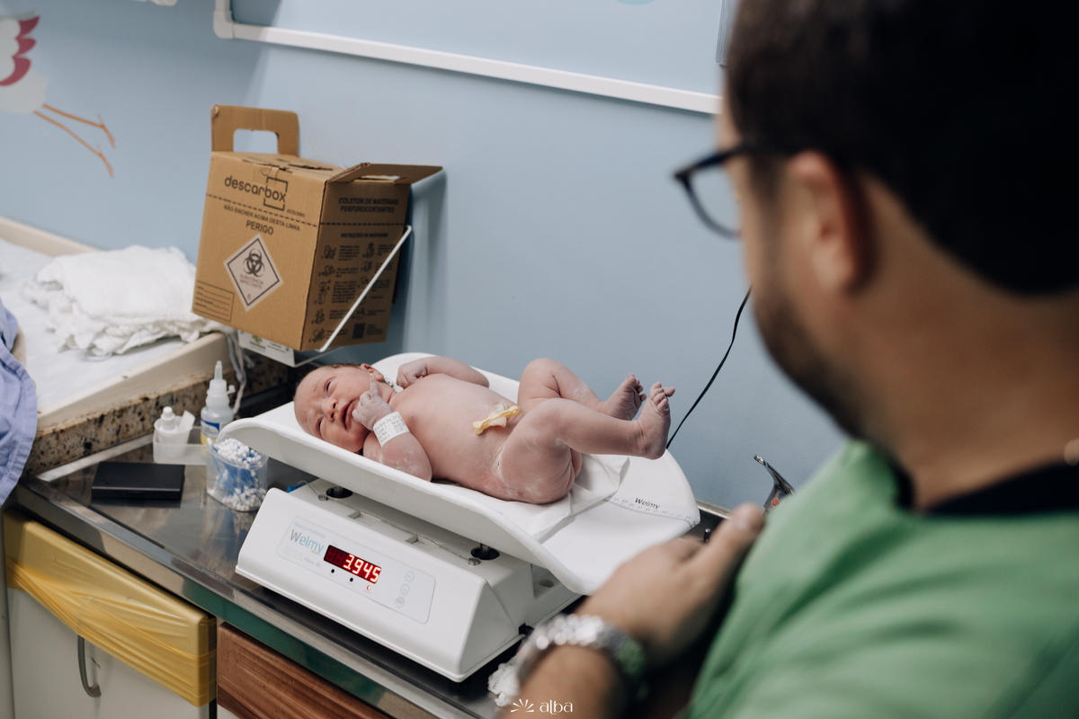 Amália recém-nascida sendo pesada na balança do Hospital Bruno Born, em Lajeado/RS, com o pai ao lado — foto feita pelo Alba Estúdio.