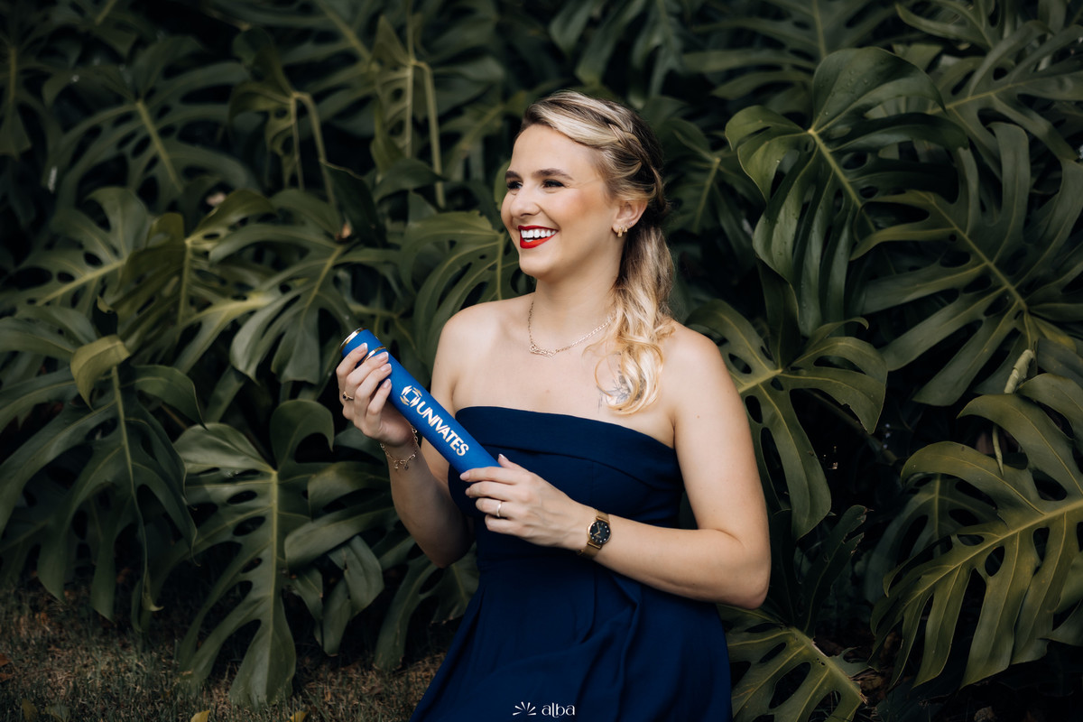 Samara em ensaio de formatura na Univates, em Lajeado, segurando o canudo azul em meio às folhagens, pelo Alba Estúdio.