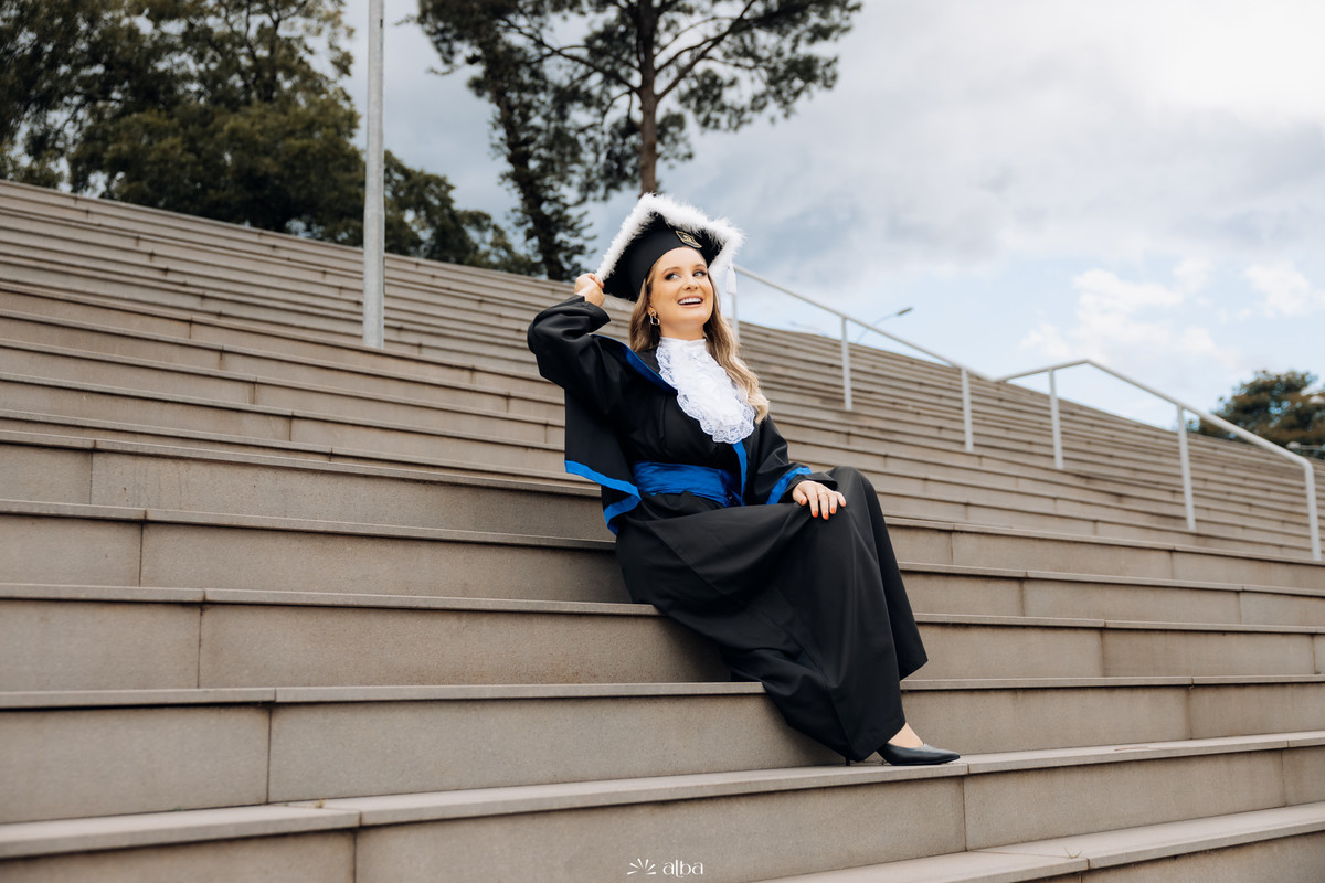 Rafaela em ensaio de formatura na Univates, em Lajeado, sentada na escadaria usando beca e capelo, pelo Alba Estúdio.