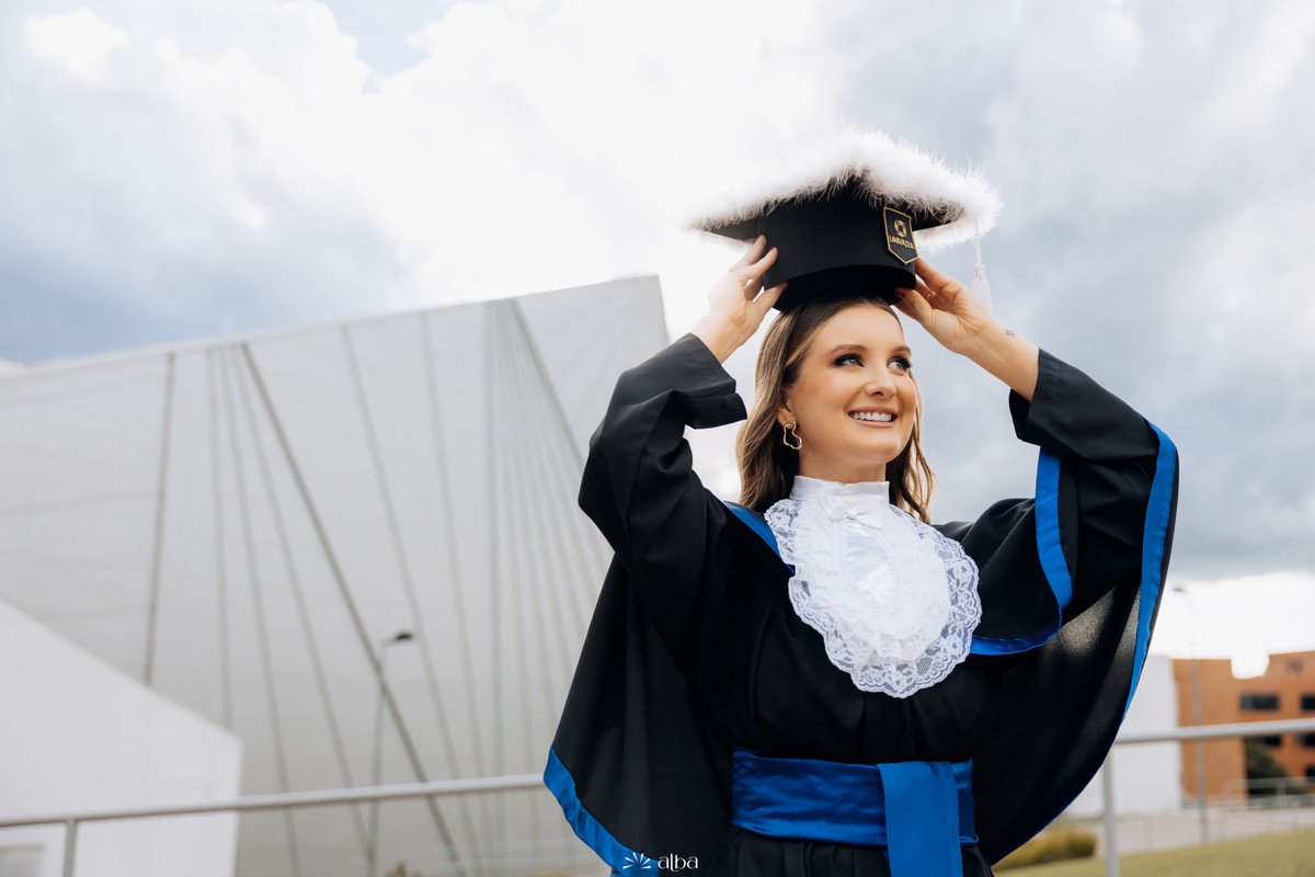 Ensaio de formatura da Rafaela na Univates, em Lajeado, com beca e capelo em frente à arquitetura do campus, pelo Alba Estúdio.