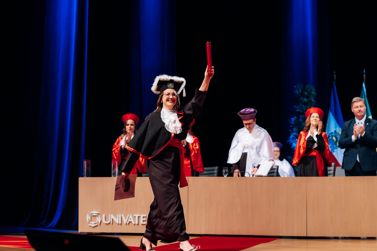 Milena celebrando sua formatura na Univates, em Lajeado, segurando o canudo no palco, registrada pelo Alba Estúdio.