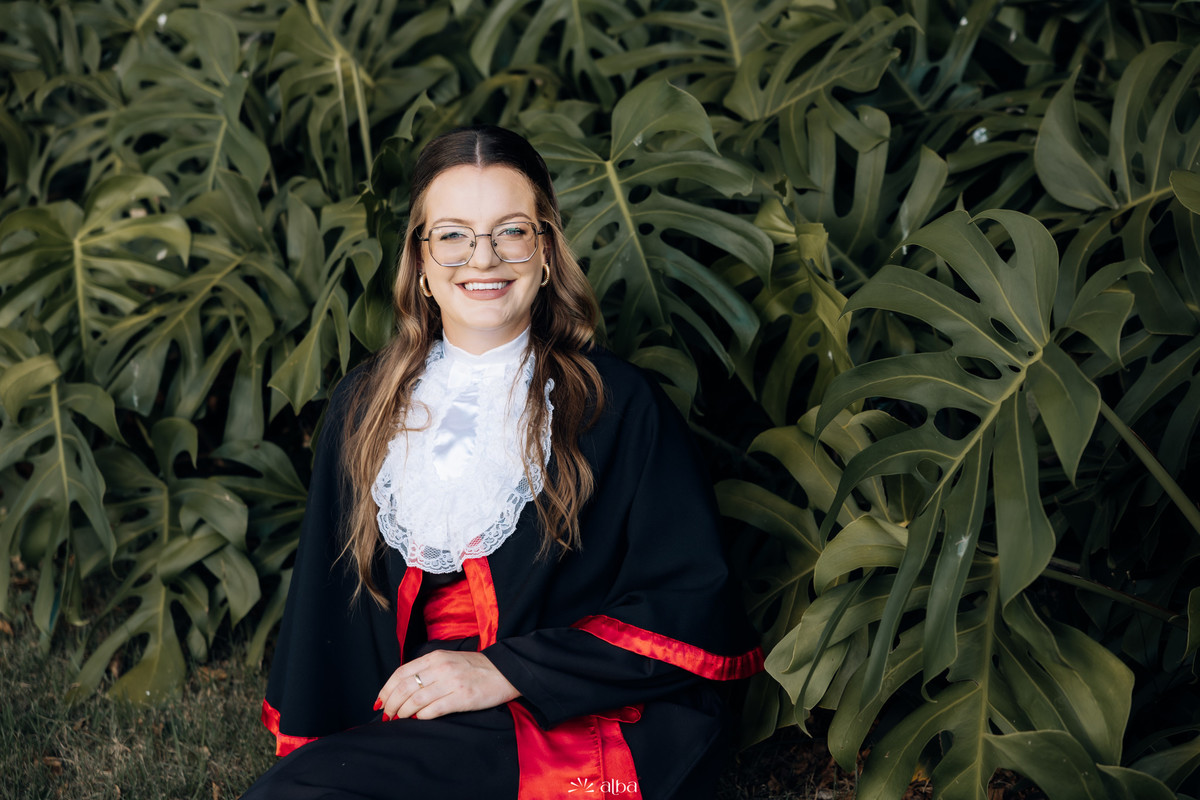 Milena sorrindo em ensaio de formatura na Univates, em Lajeado, sentada entre folhagens, fotografada pelo Alba Estúdio.
