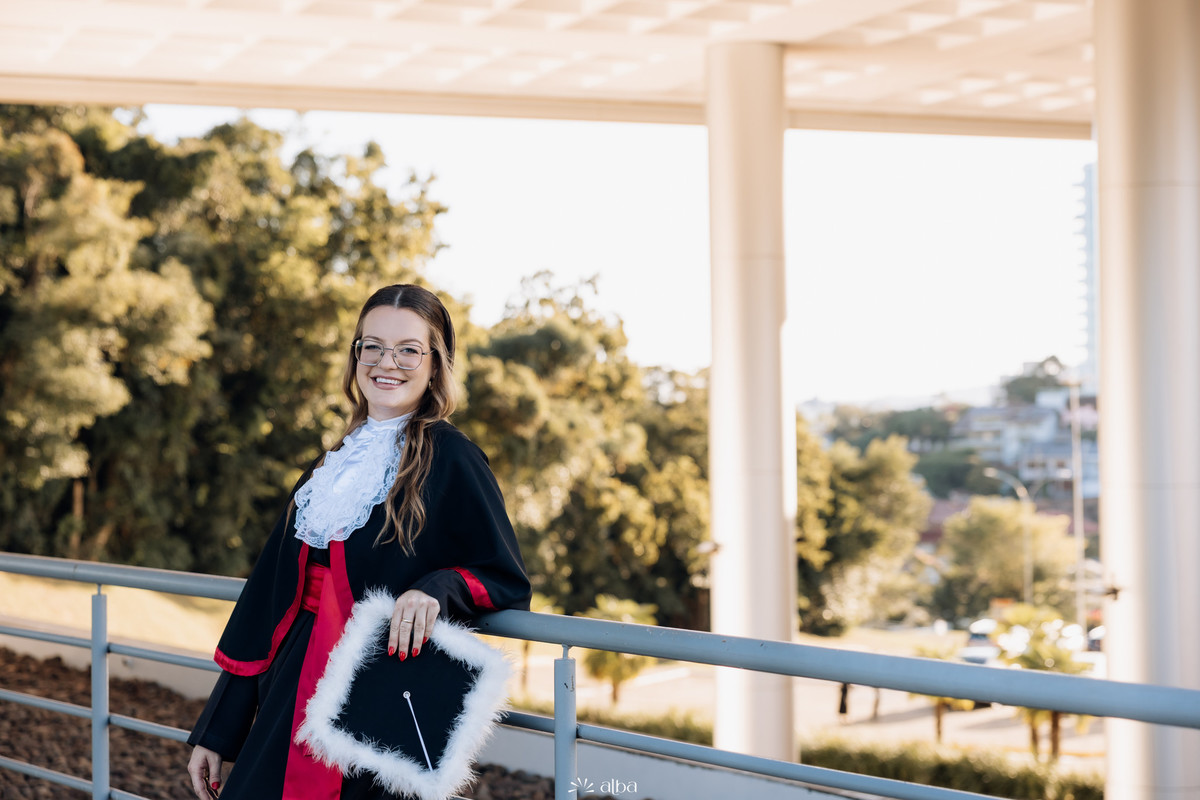 Milena em ensaio de formatura na Univates, em Lajeado, usando beca e segurando o capelo, fotografada pelo Alba Estúdio.