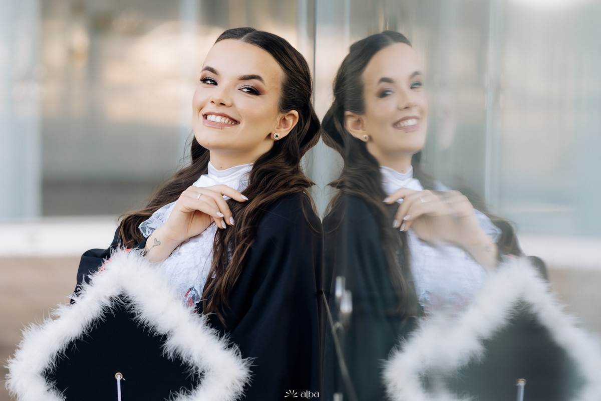 Retrato da formanda Alice na Univates, em Lajeado, com beca, capelo e reflexo no vidro, pelo Alba Estúdio.