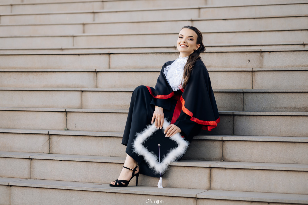 Alice sentada nas escadarias da Univates, em Lajeado, durante ensaio de formatura com beca e capelo, pelo Alba Estúdio.