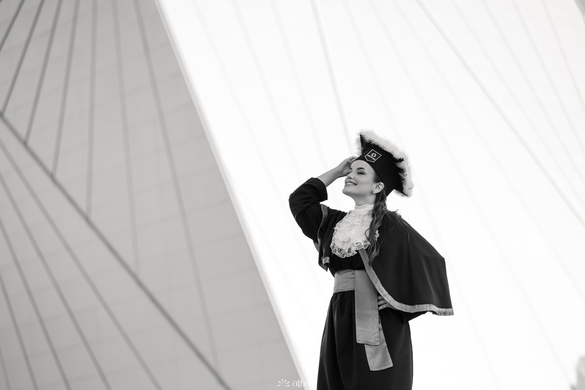 Alice em foto preto e branco durante ensaio de formatura na Univates, em Lajeado, com capelo e arquitetura ao fundo, pelo Alba Estúdio.