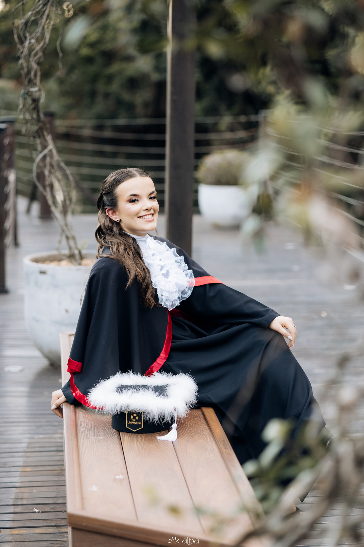 Alice em retrato de formatura na Univates, em Lajeado, sentada em banco externo com beca e capelo, pelo Alba Estúdio.