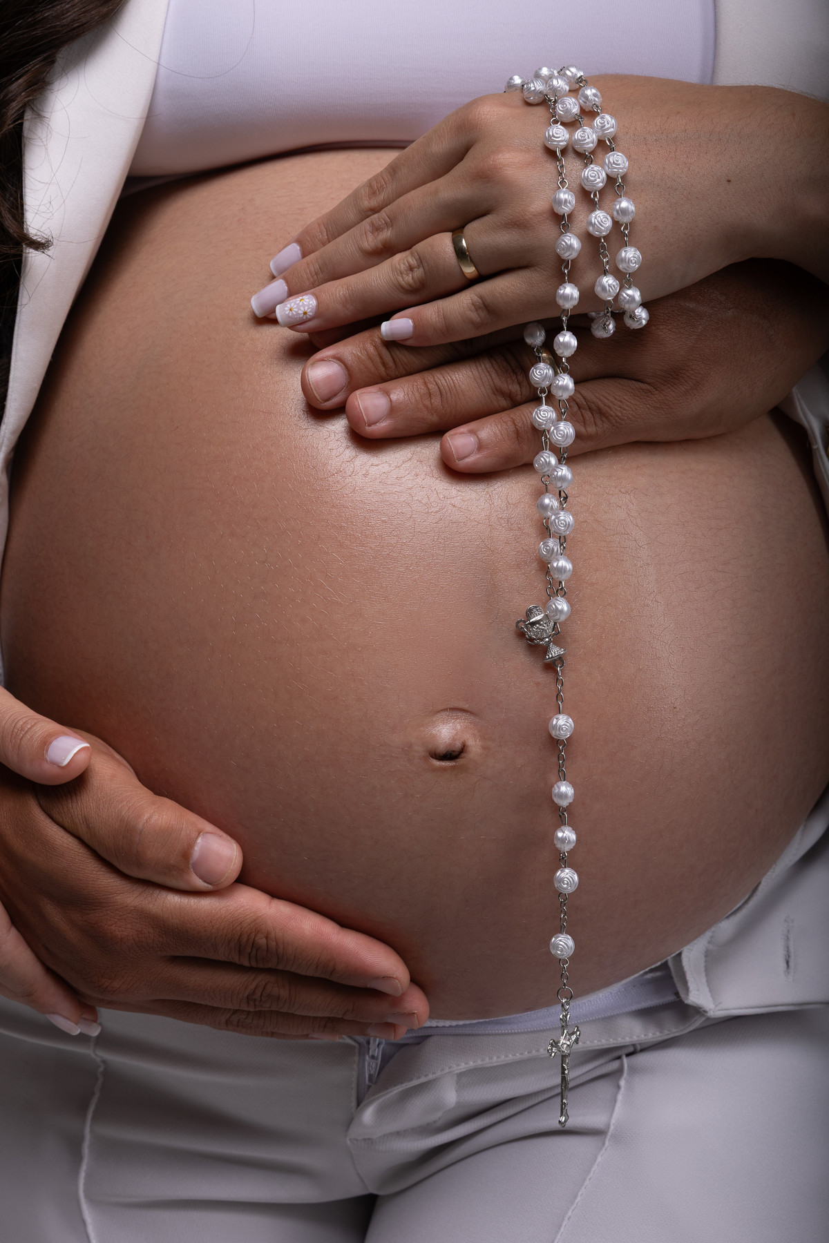 Detalhe da barriga de gestante com as mãos do casal e um terço, representando fé, amor e espera. Ensaio fotográfico do Alba Estúdio em Lajeado, RS.