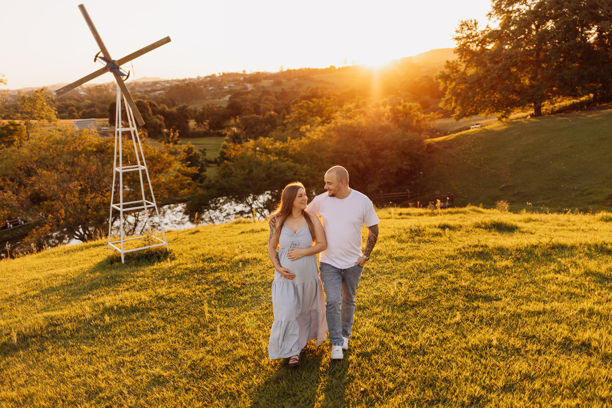 Casal caminhando durante ensaio gestante externo ao pôr do sol, em cenário de campo com moinho | Alba Estúdio, Lajeado RS.
