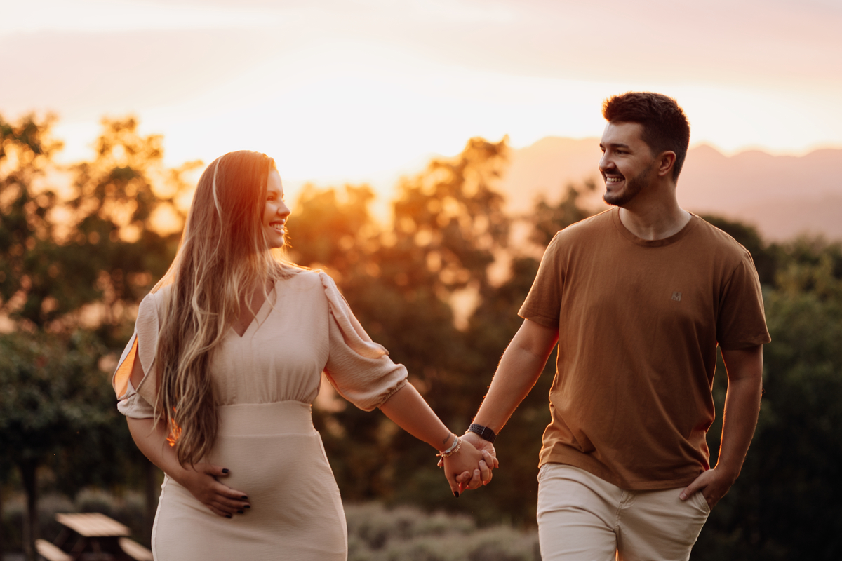 Casal caminhando de mãos dadas ao pôr do sol durante ensaio gestante externo | Fotografia de gravidez pelo Alba Estúdio.