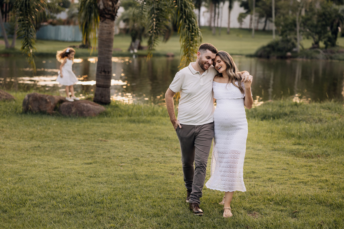 Casal gestante caminhando próximo ao lago enquanto a filha brinca ao fundo | Ensaio de família em Lajeado pelo Alba Estúdio.