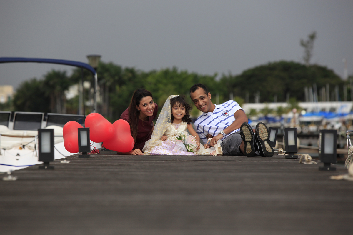 foto tirada na beira do lago paranoa com fundo de cenario barcos e outras decoracoes, nico pode capturar esse momento onde papai, mamae e filha posam.