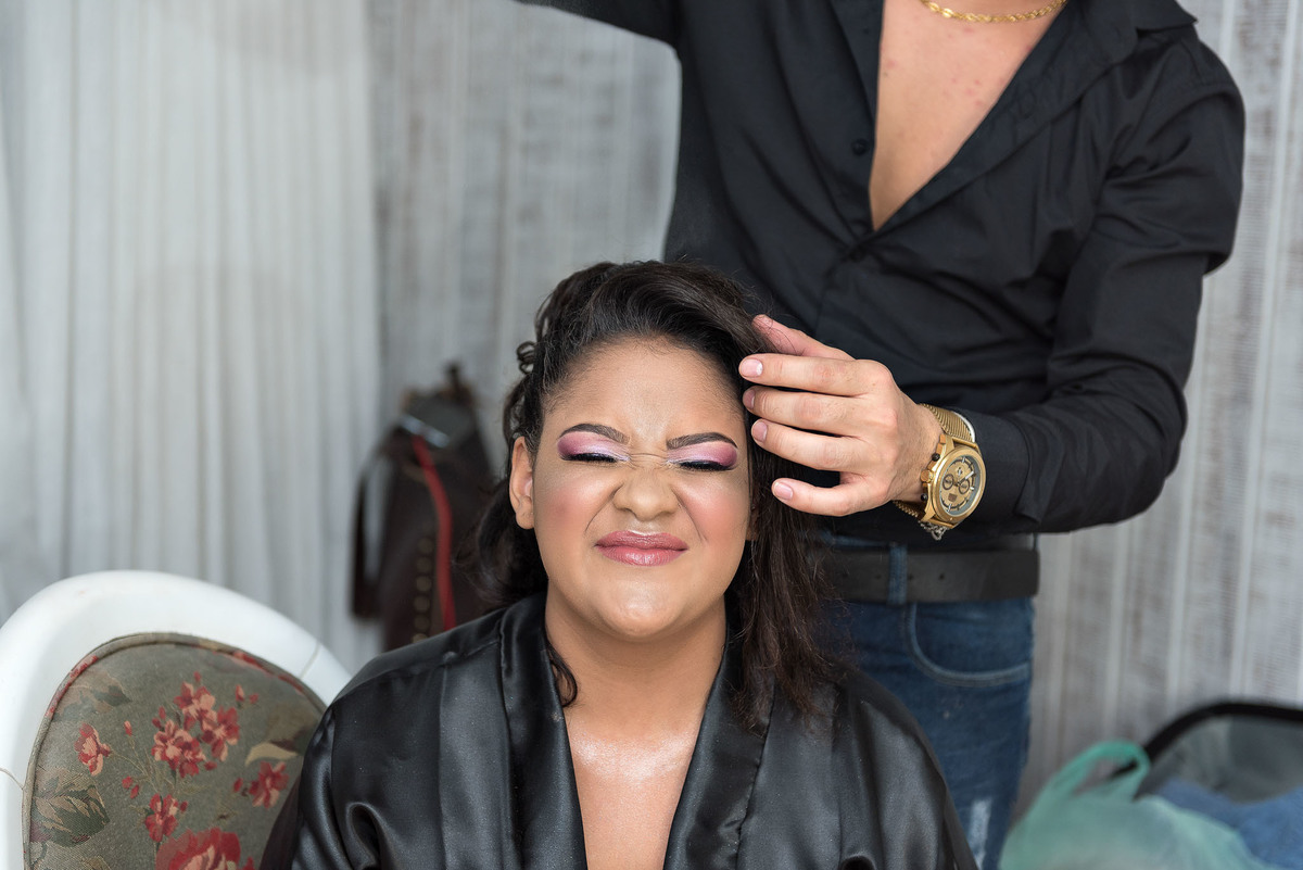 Jamile, a debutante, sorrindo enquanto faz maquiagem e cabelo, com maquiadores ao redor, em um momento de preparação para a festa