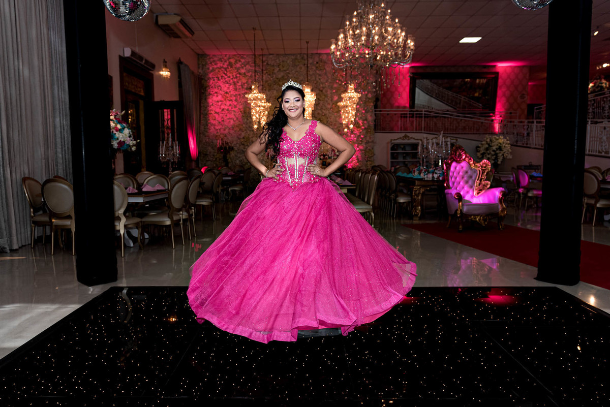 Jamile em seu vestido de debutante rosa choque, posando majestosamente em frente à entrada do salão de festas, com decoração iluminada