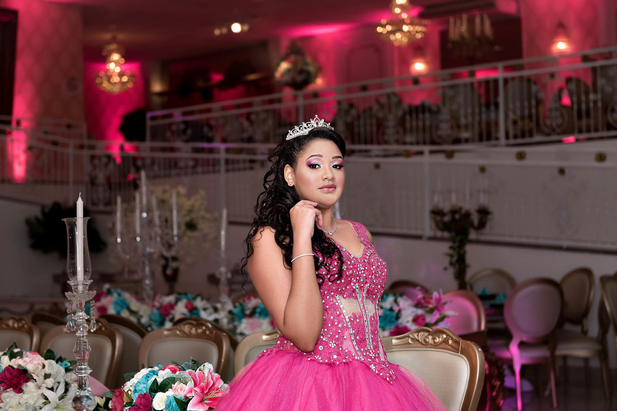 Jamile em pose clássica de debutante, com o vestido rosa choque em destaque, e o ambiente do salão de festas como fundo, no Marilias Buffet