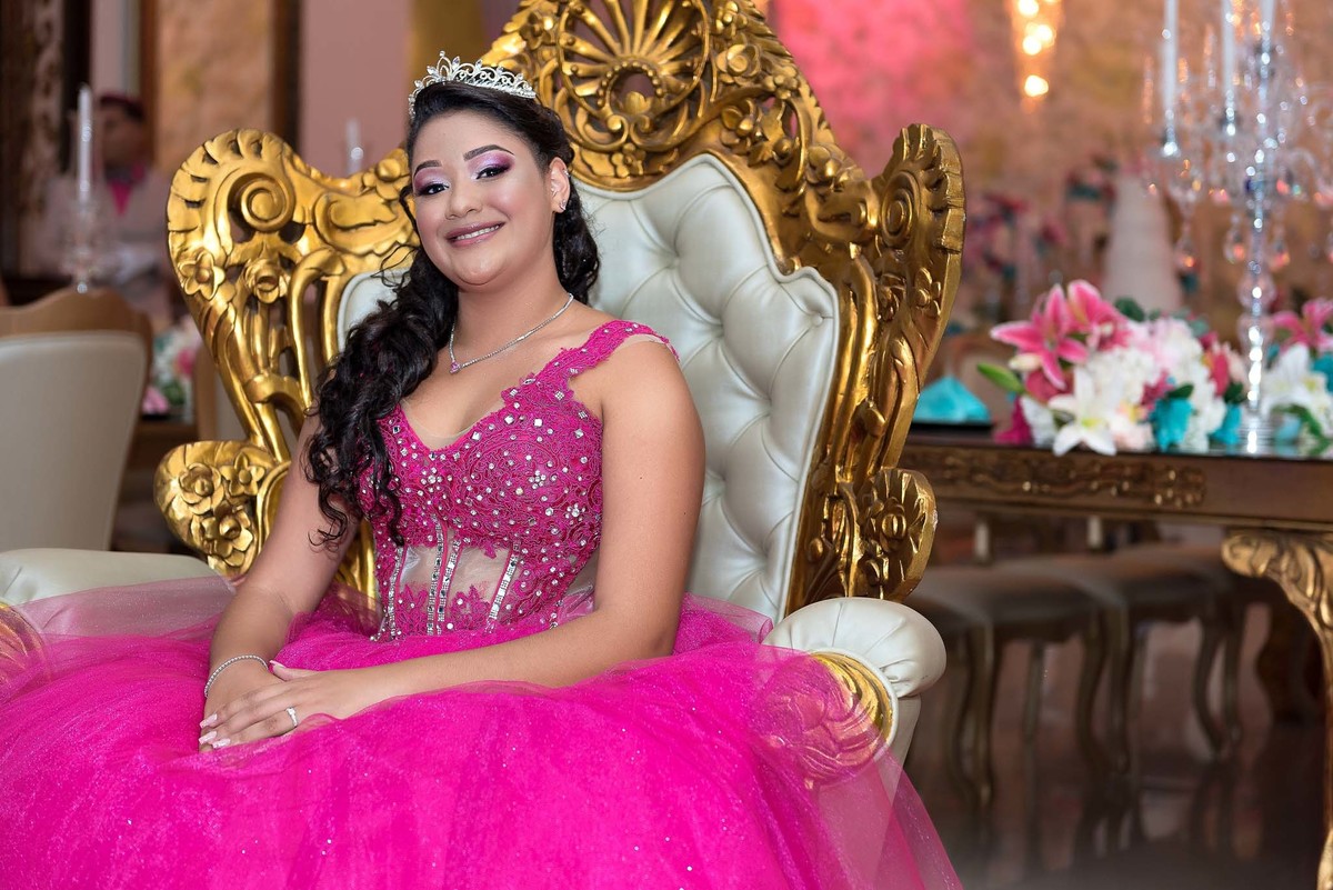 Jamile posando no trono dourado, olhando para a câmera e sorrindo, com detalhes da decoração ao fundo