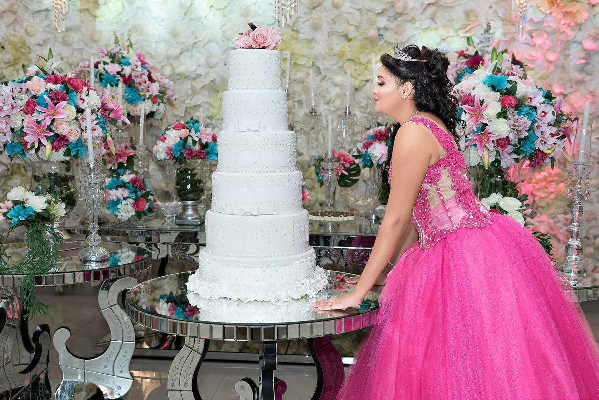 Jamile, a debutante, em seu vestido rosa choque, ao lado de um bolo de 15 anos branco e alto, decorado com flores rosas