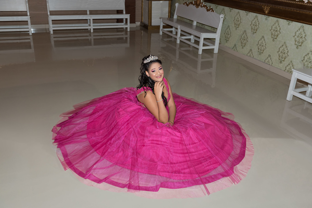 Jamile, em seu vestido rosa choque vibrante, deitada em um sofá de veludo branco, em uma pose descontraída