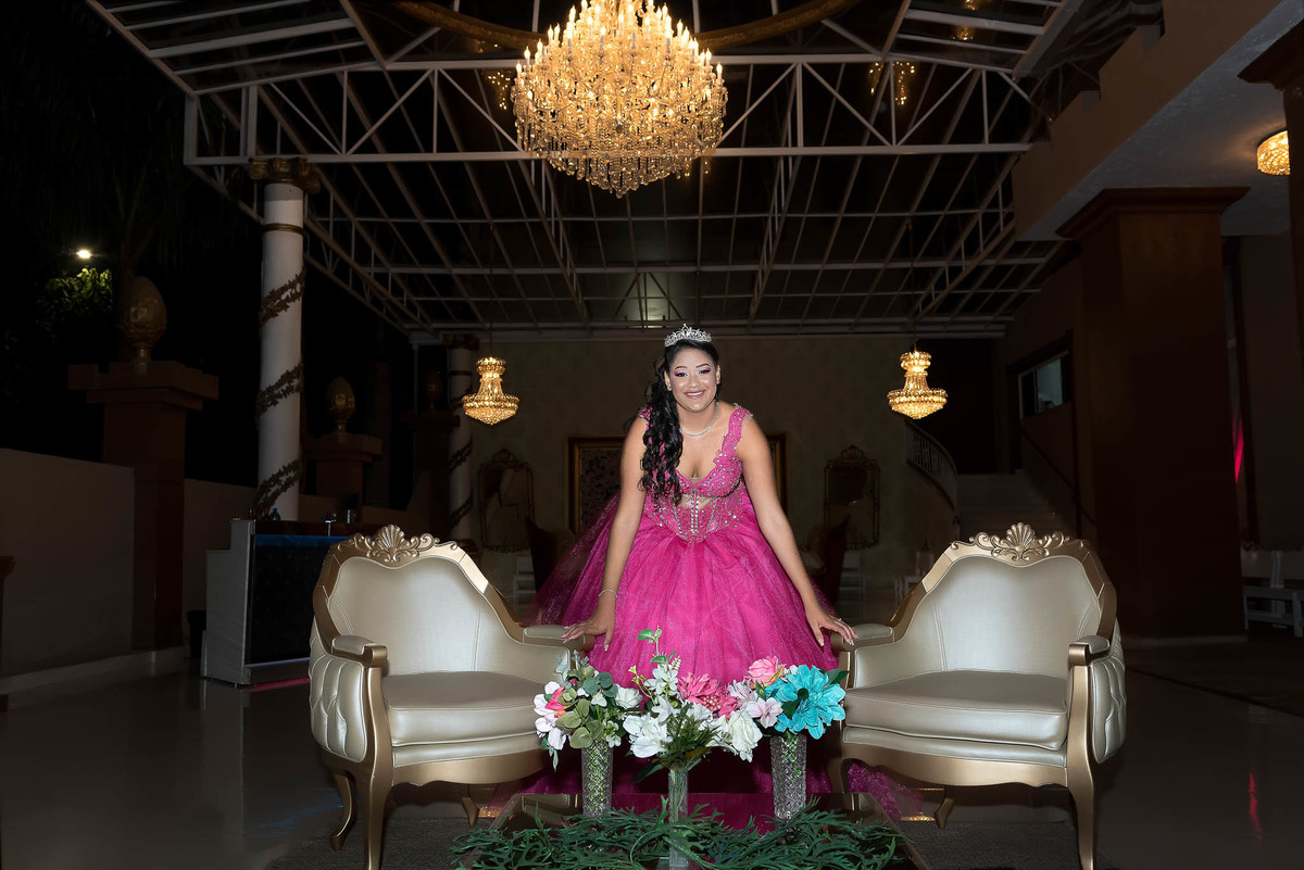 Jamile, a debutante, em seu vestido rosa choque, sentada em uma mesa de festa com cadeiras luxuosas ao fundo, dentro do salão
