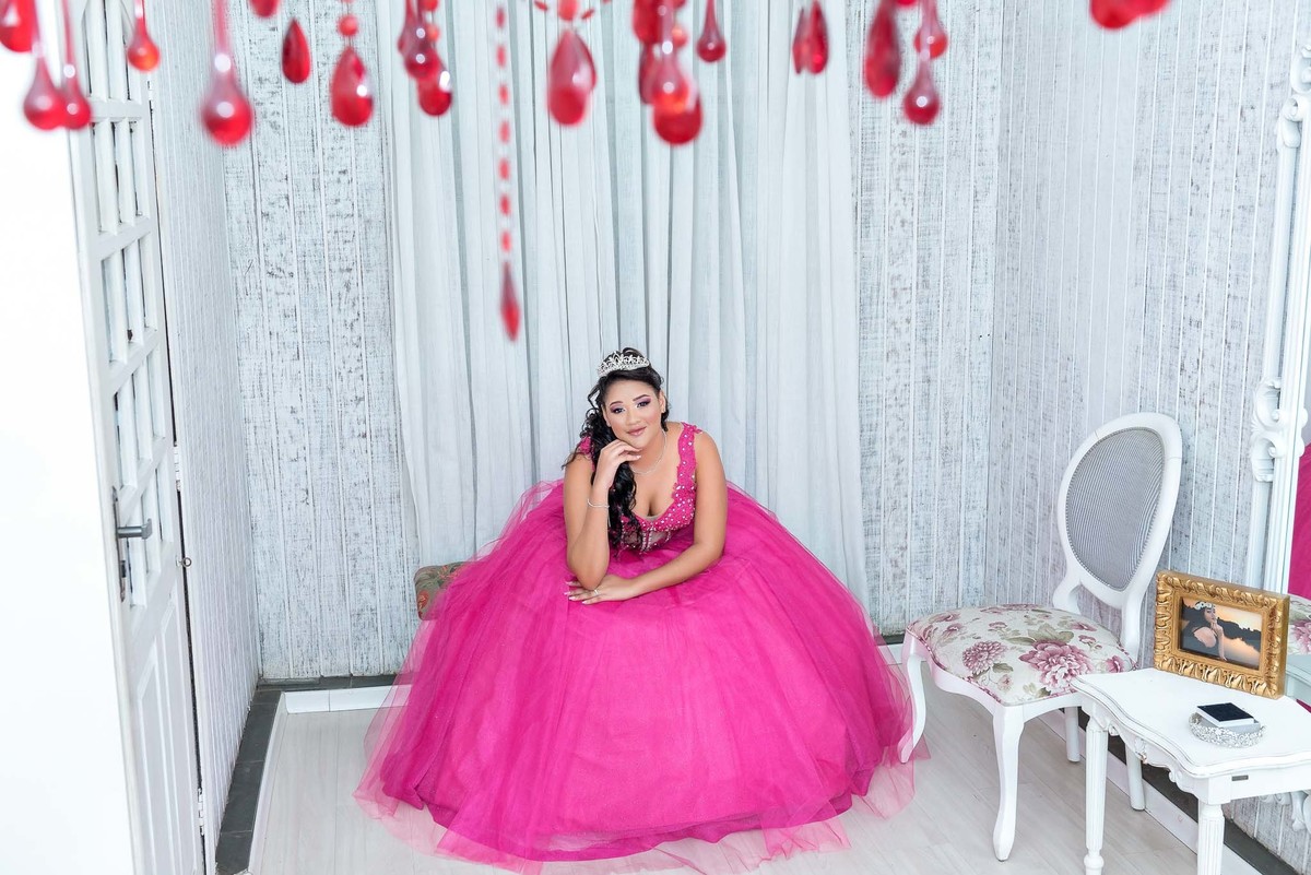 Jamile em seu vestido rosa choque, sentada em uma cadeira branca adornada com flores, com iluminação em tons de rosa