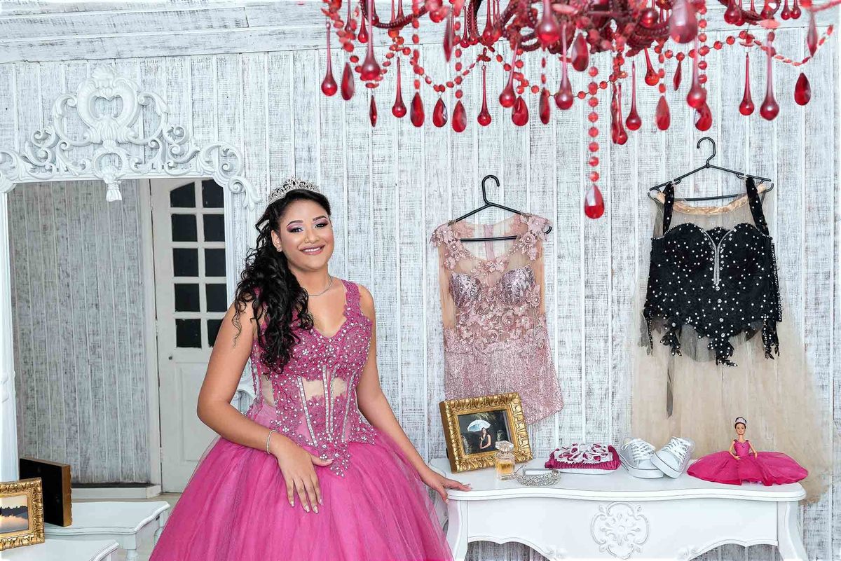Jamile em seu vestido rosa choque, posando em frente a um closet aberto exibindo vestidos e acessórios, mostrando um momento de escolha de figurino