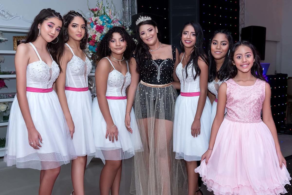 Jamile e suas amigas em vestidos de festa, algumas de branco e outras de rosa e lilás, posando juntas, celebrando a festa de 15 anos