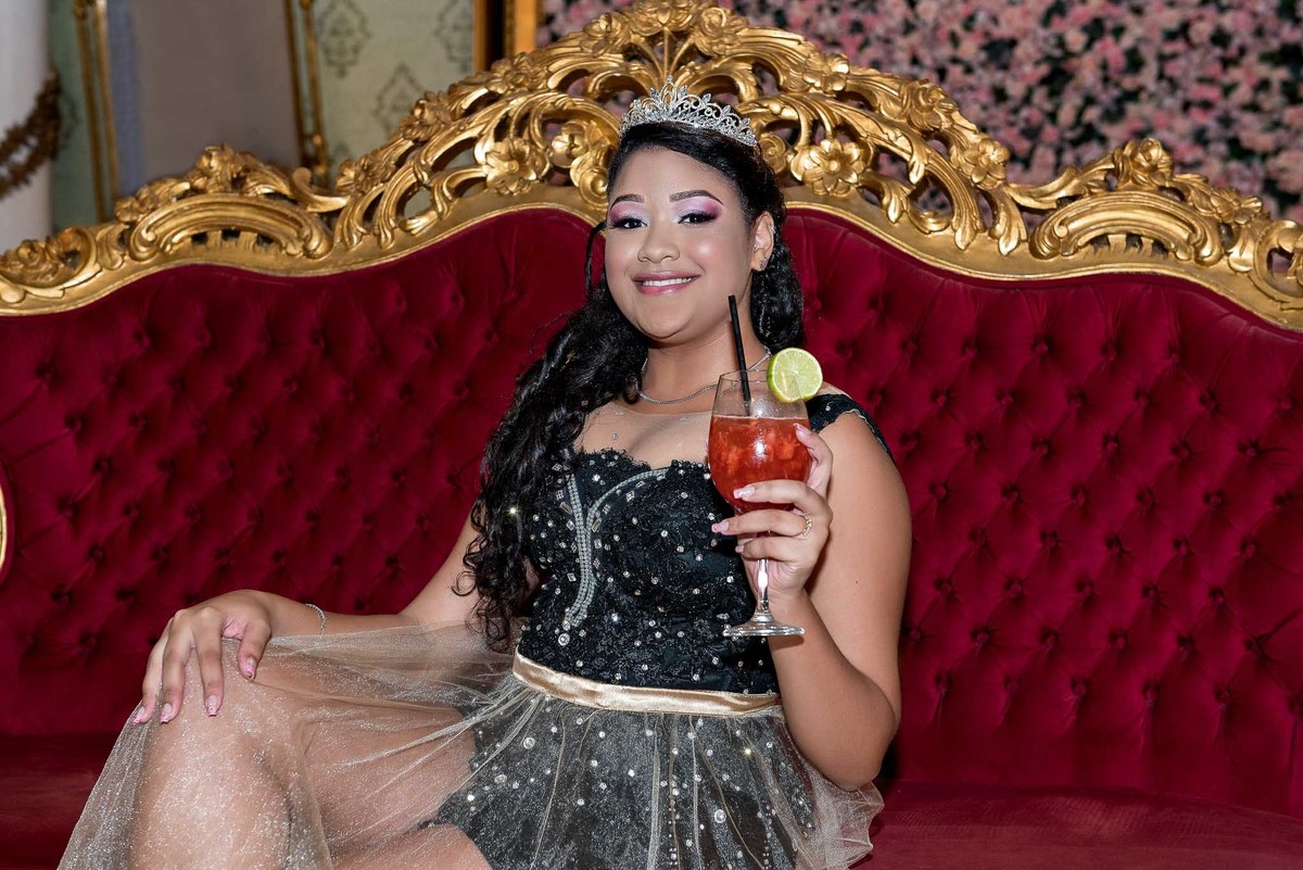 Jamile em um vestido preto e brilhante, sentada em um sofá de veludo vermelho, com um drink na mão, em um ambiente mais sofisticado