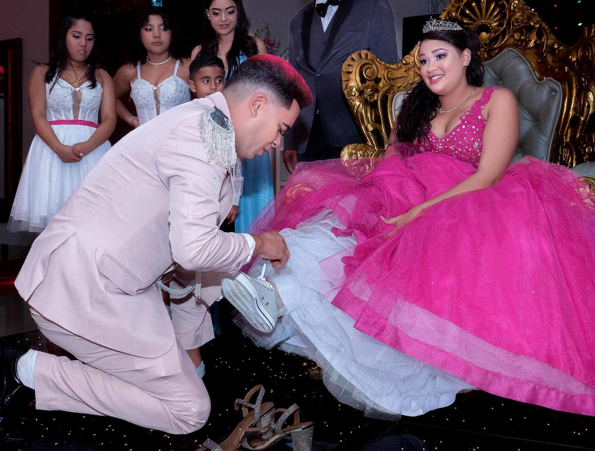 Príncipe colocando o sapato e a vista dos convidados da festa de 15 anos, incluindo adultos e jovens, sentados em mesas ou assistindo à celebração