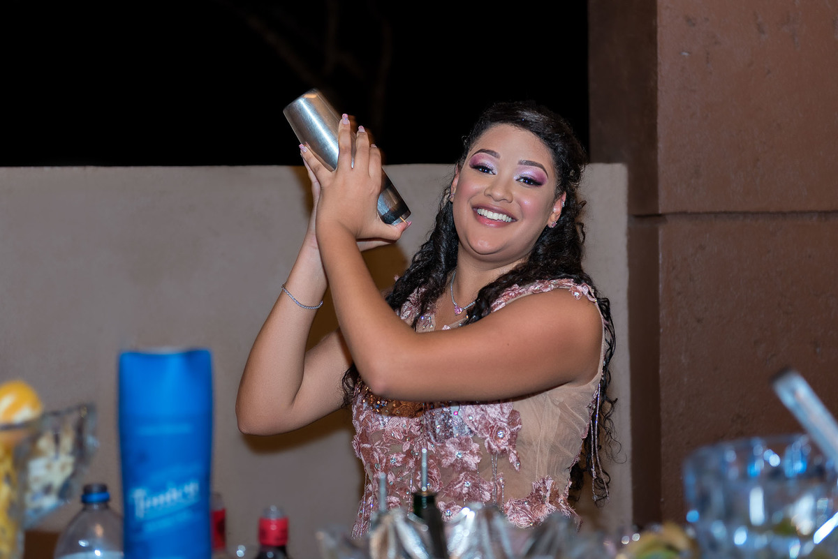 Jamile em um vestido rosa brilhante, no bar da festa, fazendo pose e misturando uma bebida, como uma bartender