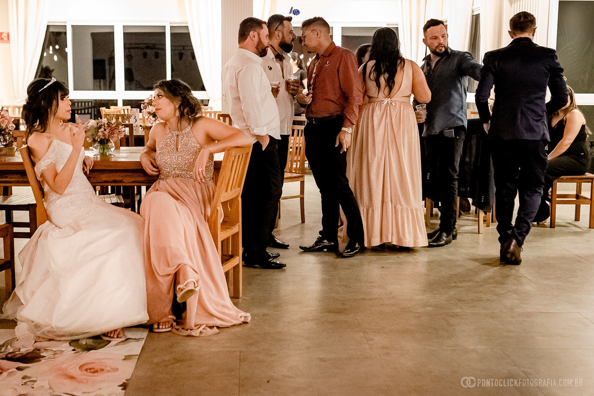 Noiva sentada com a madrinha conversando grupo de pessoas em pé conversando ao fundo